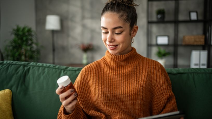 A woman sits on a couch considering a bottle of pills.