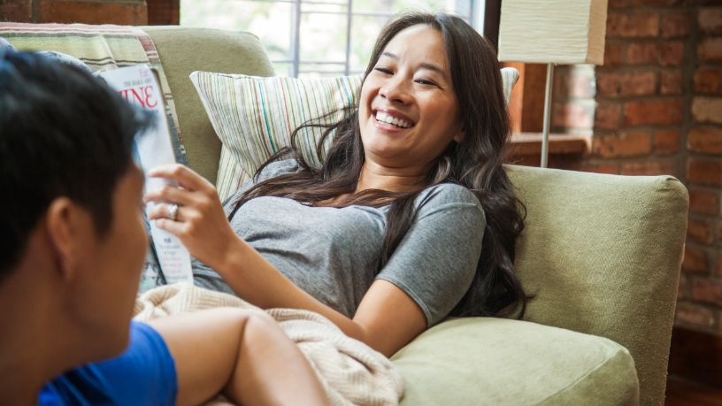 A woman lays on a sofa talking to a man nearby.