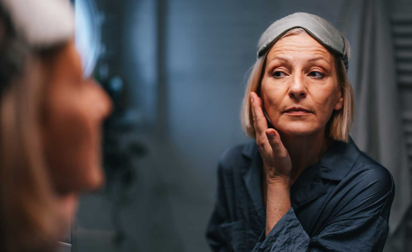 Woman wearing sleep mask examining her face in the mirror