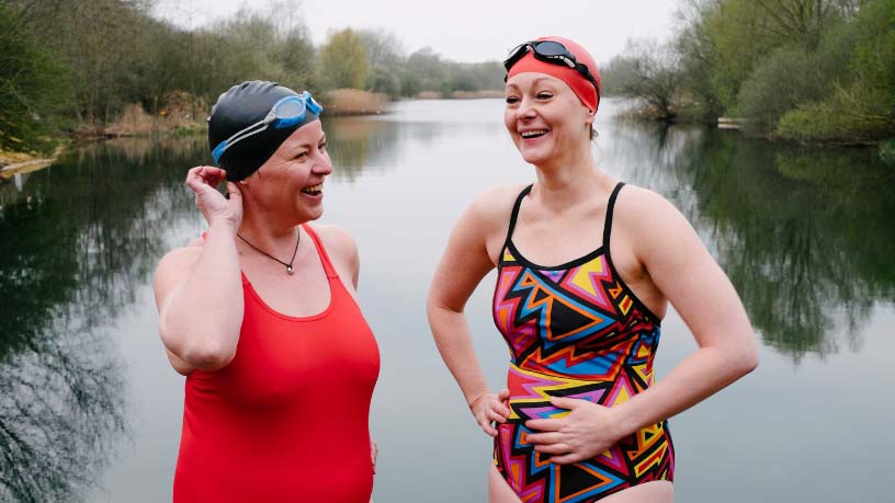 Two women in bathers stand in front of a lake and laugh.