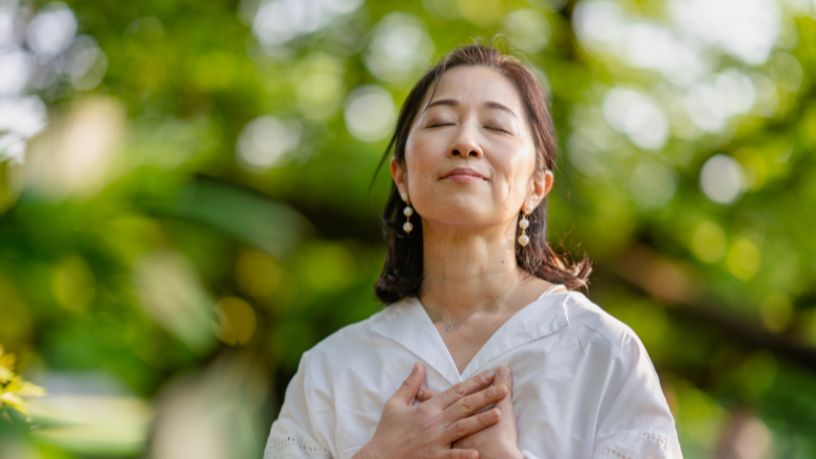 A woman stands outside with her eyes closed and hands pressed to her chest as she breathes.