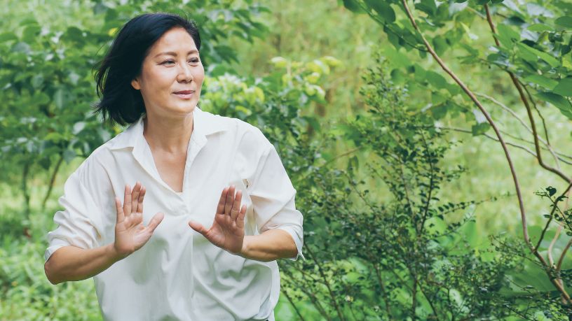 A woman stands outside, moving through a qigong flow with her hands in front of her chest to push energy from her body.