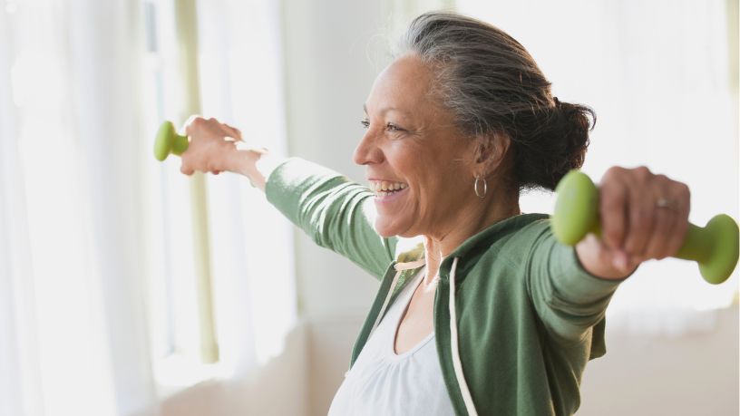 A woman smiles while holding weights with her arms out straight.