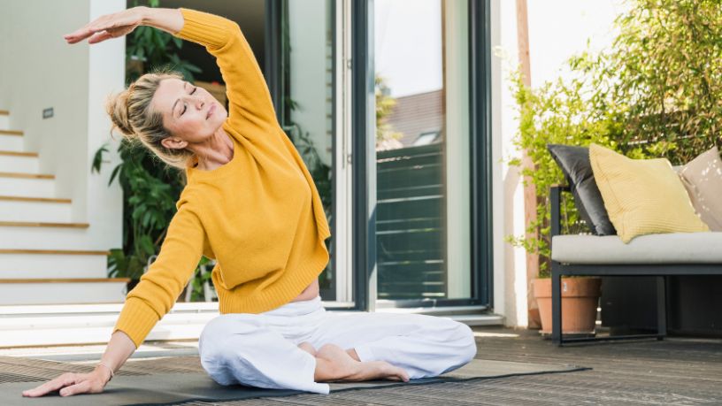 A woman sits on a yoga mat with her legs crossed, leans to the side and stretches her arm over her head.