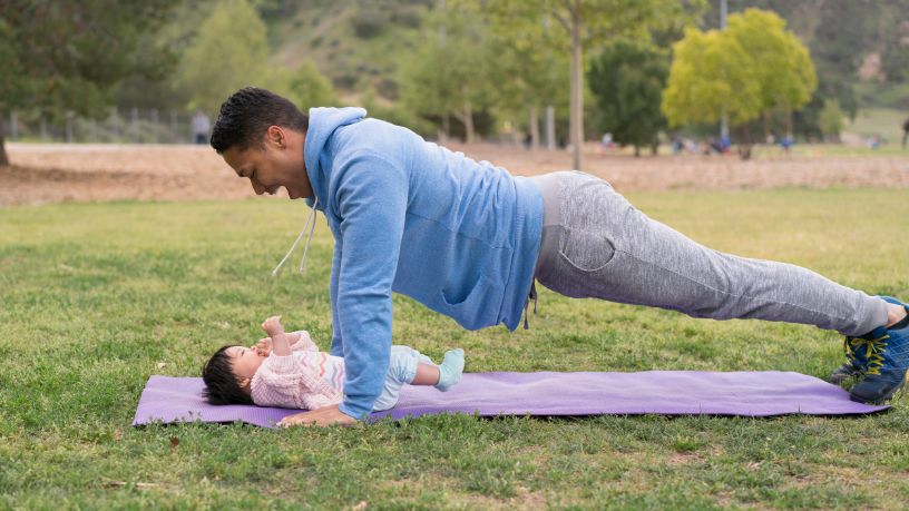 A man holds a plank and smiles down at his baby, who lies on the yoga mat below him.
