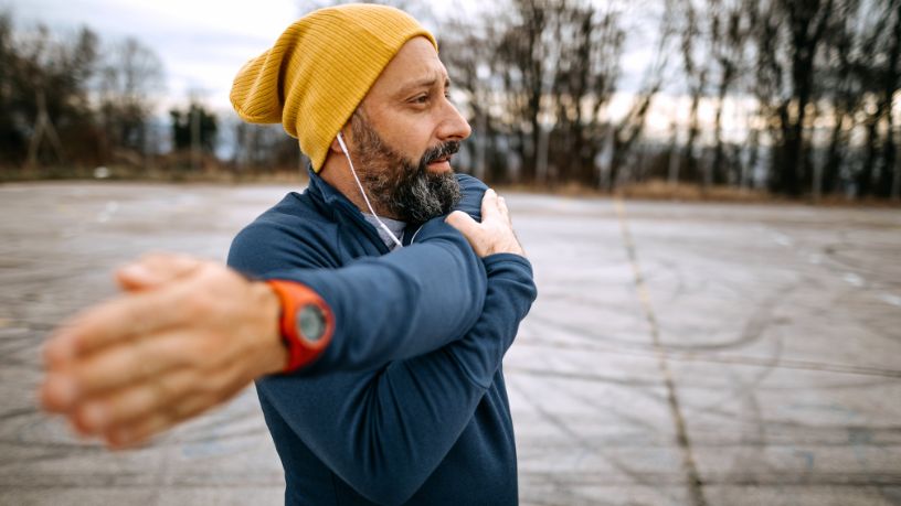A man stands outside as the sun comes up, stretching his left arm across his body and getting ready for a run.