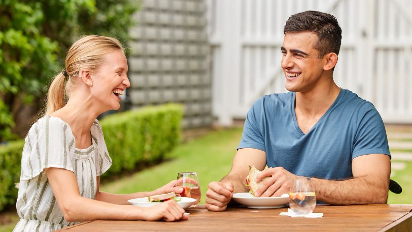 Two people sit at an outdoor table, laughing and eating sandwiches.