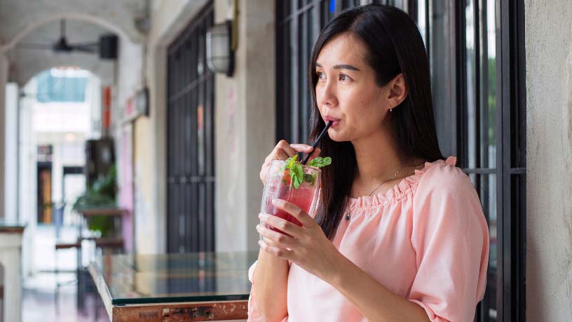 A woman stands outside a bar drinking a pink cocktail.