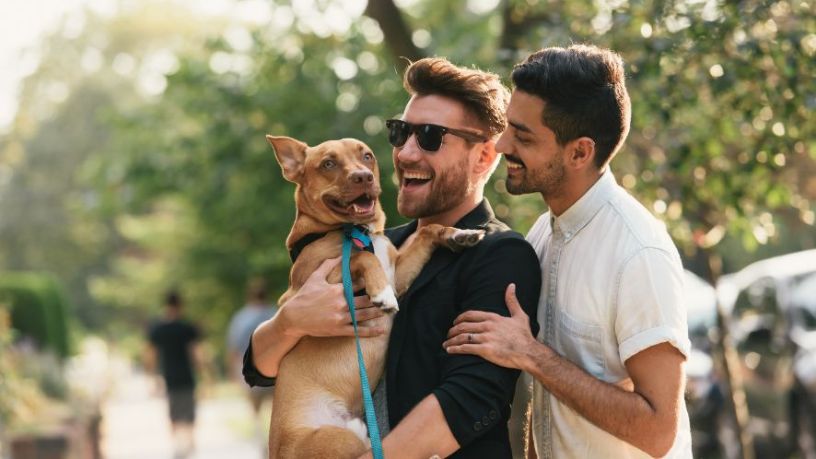 Two men smile and hold a dog in a park.