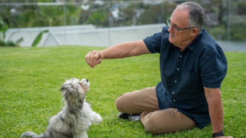Man sitting on lawn holding up a treat for a little puppy.