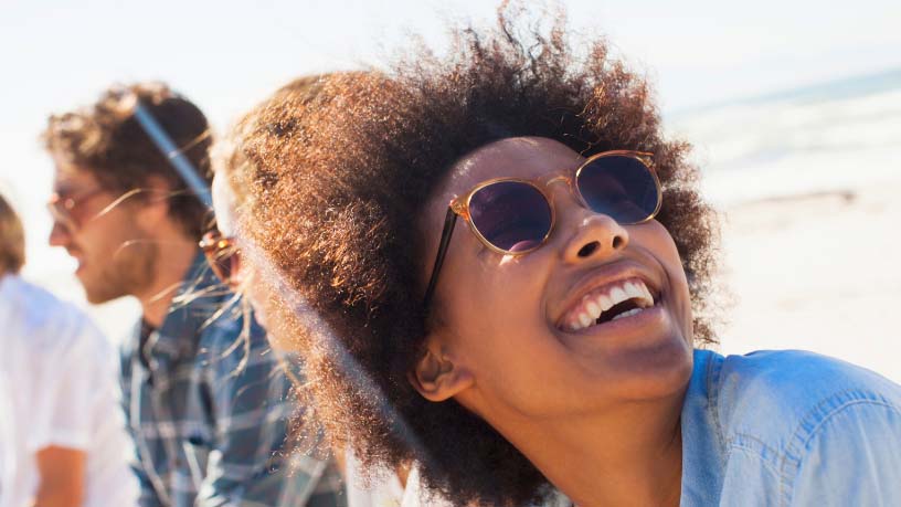 A woman wearing sunglasses at the beach grins at the sky.