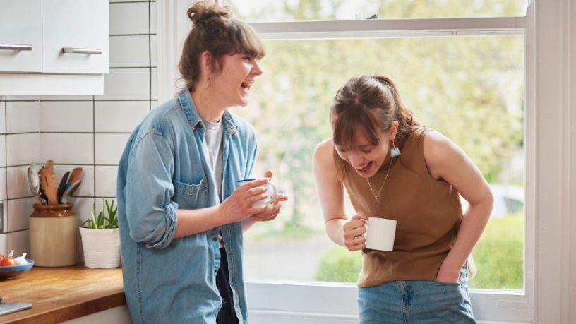 Two young women laughing in the kitchen holding coffee cups.