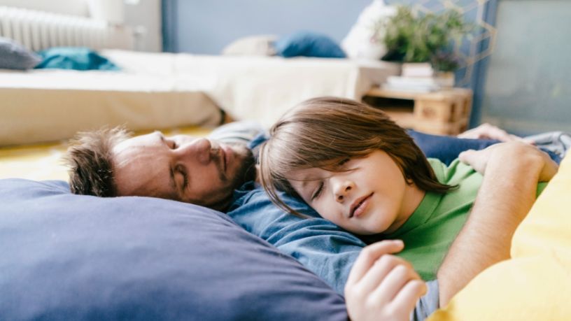 A man and his son sleep next to each other on the living room floor.