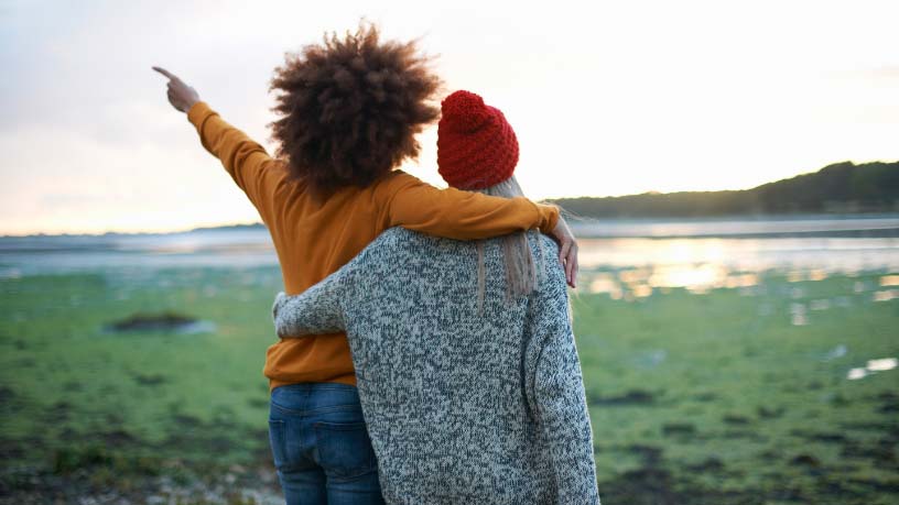 Two women stand by a river with their arms around each other looking at a sunrise.