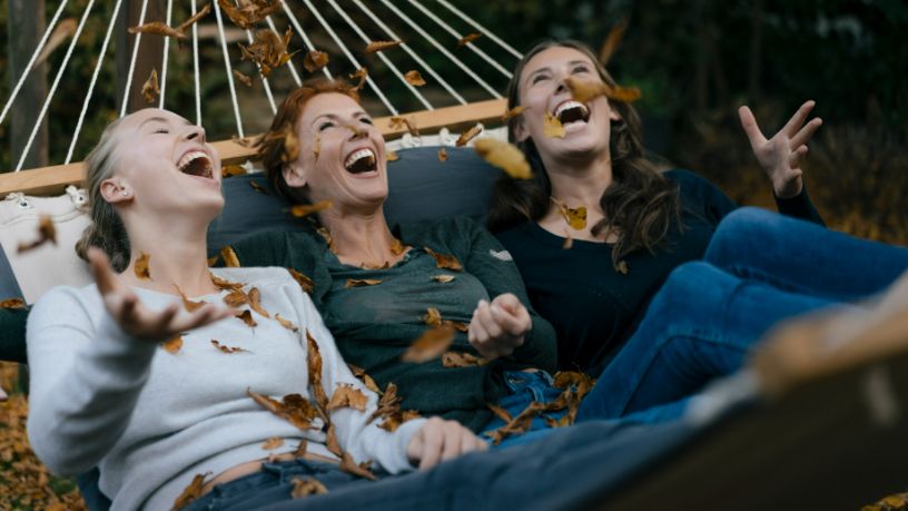 Three women in a hammock laugh as leaves fall from a tree.