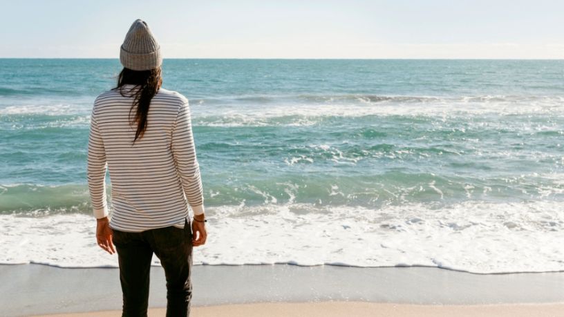 A person wearing a beanie stands on the beach looking out over the ocean.