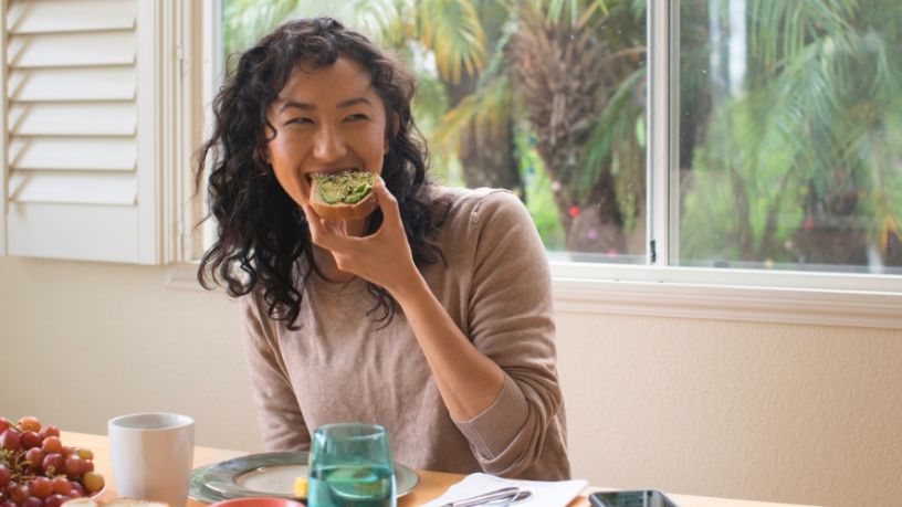 A woman sits at a dining table and eats avocado on toast.