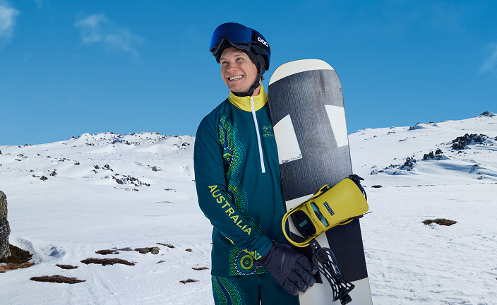 Athlete Ben Tudhope in green and yellow Australian uniform standing on snow holding a snowboard. Mountain landscape behind.