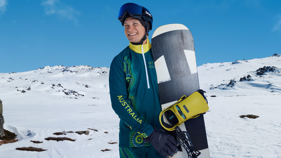 Athlete Ben Tudhope in green and yellow Australian uniform standing on snow holding a snowboard. Mountain landscape behind.