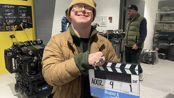 A person in a beanie and brown jacket smiling on a film set while holding a movie clapperboard.