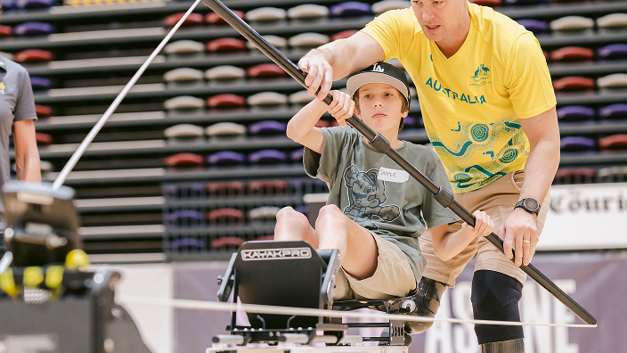 Adult in yellow Paralympian shirt assisting a child seated in adaptive rowing equipment inside a stadium setting.