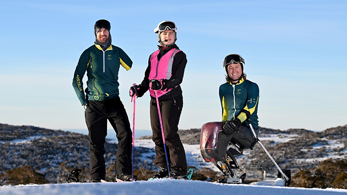 Three Para athletes on a snowy mountain. One standing, one seated using adaptive ski equipment, one in winter gear. Snow landscape.