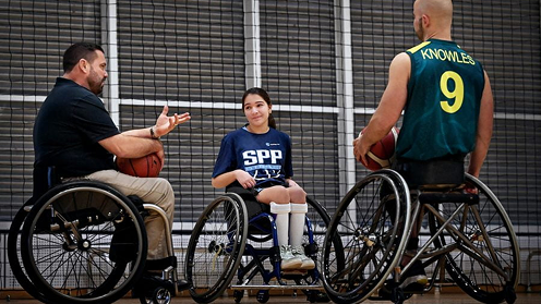 A basketball coach talking to a young girl and an adult athlete, all using wheelchairs, on an indoor court.