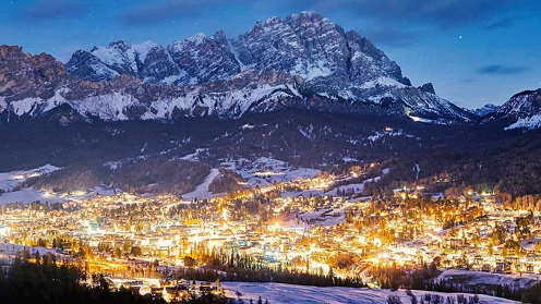 A wide, scenic night view of a glowing alpine village at the base of snow-capped mountains in Milano Cortina.