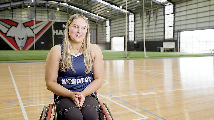 Harriet sitting in a sports wheelchair on an indoor basketball court, smiling warmly at the camera.