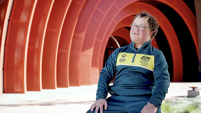 James wearing a green and gold Australian team jacket, sitting outdoors in front of a large, curved orange architectural structure.