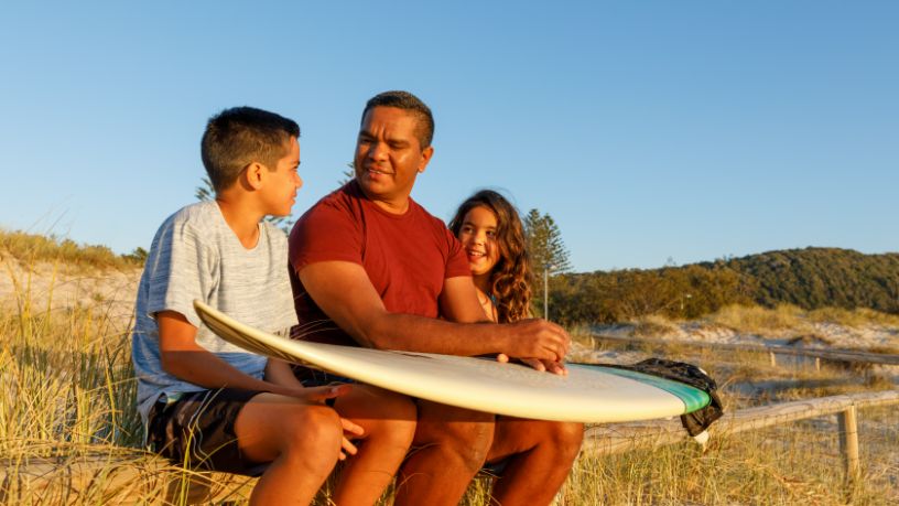 A man with a surfboard rested on his lap sits between his son and daughter in front of a grassy sand dune.