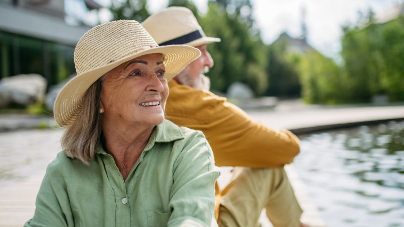 A couple wearing hats sit back to back by a pond.