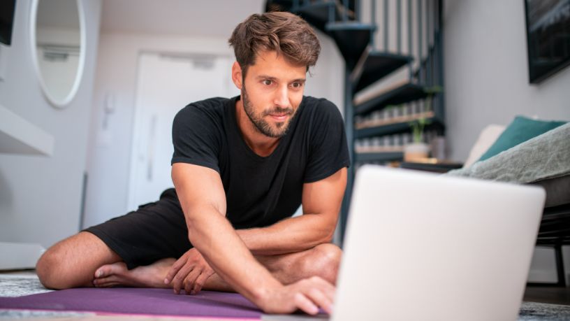 A man sits on a yoga mat in a small apartment and uses his laptop.