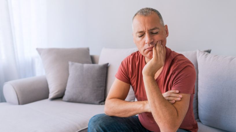 A man nurses his saw jaw as he sits on a couch.