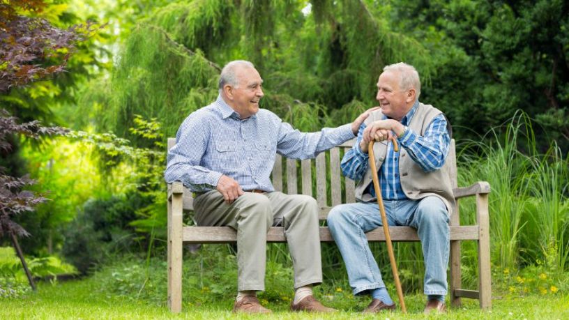 Two elderly men sit on a bench together in a park. One man holds a walking stick while the other pats him on the shoulder. 