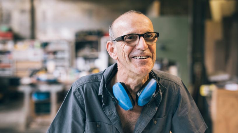 A man with a pair of earmuffs around his neck stands in a workshop.