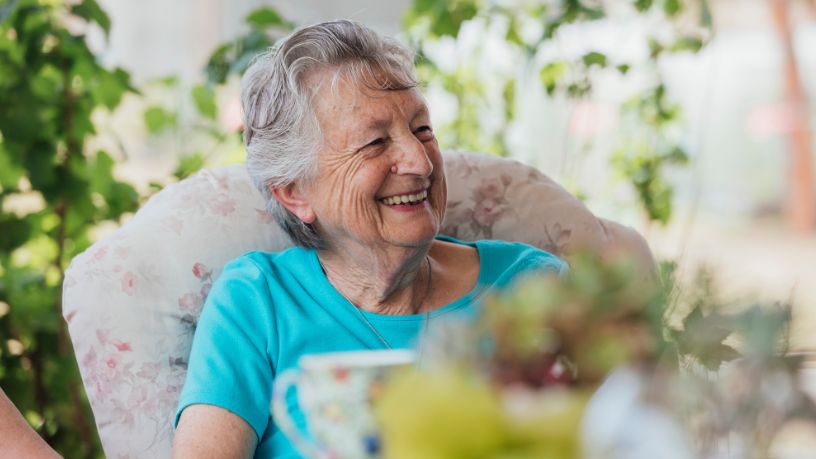 An older woman laughs while enjoying morning tea outside.