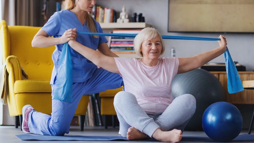 An older woman stretches a resistance band behind her head while a medical professional helps stabilise her arms.