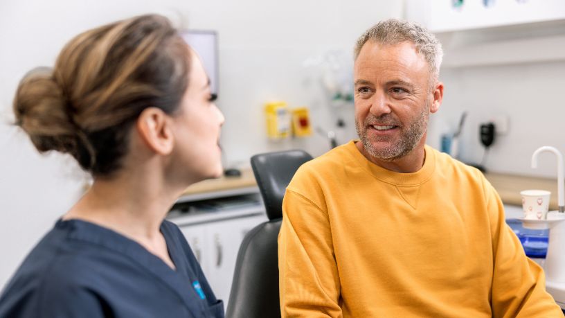 A man sits in a dentist's chair and speaks with a dentist.