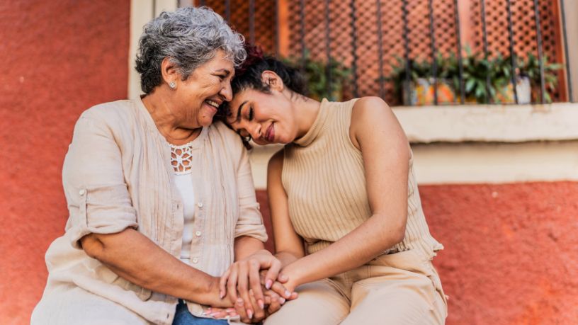A young woman rests her head on an older woman's shoulder while they hold hands and smile.