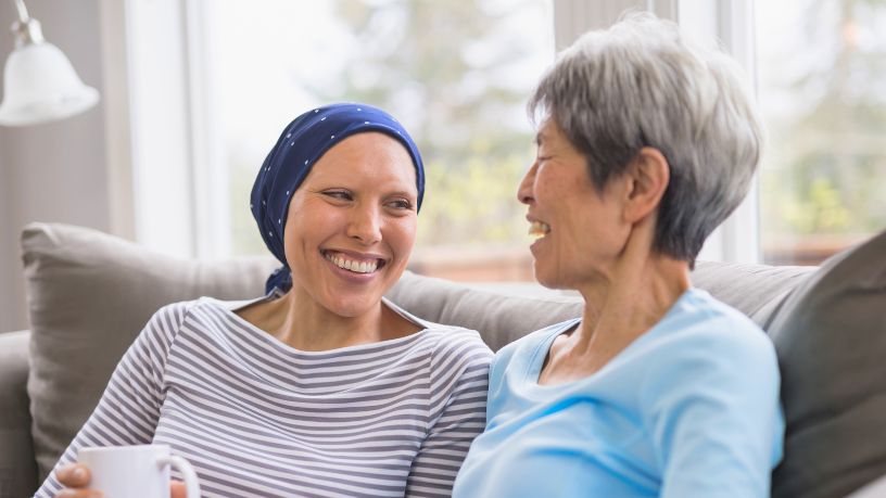 A woman wearing a headscarf smiles with her friend while they sit on a couch.