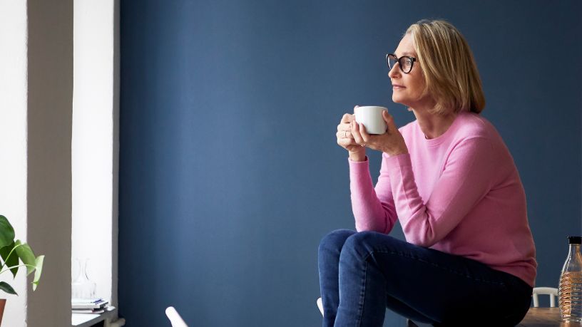 A woman sits on a table, drinking a cup of tea and looking out the window.