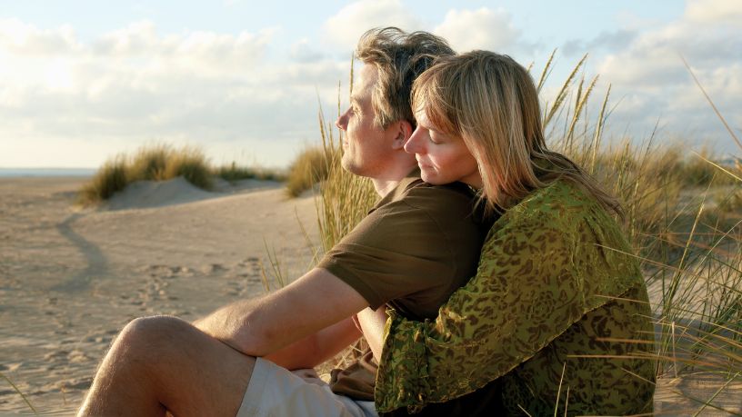 A woman sits behind a man on the beach, holding him.