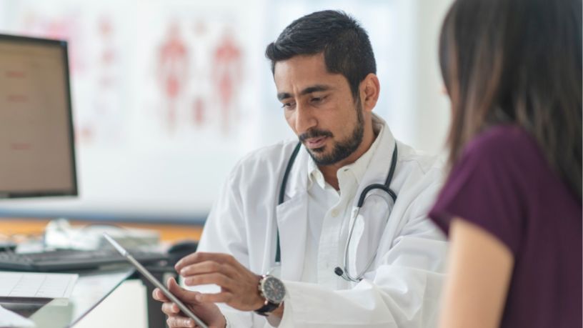 A doctor holding a tablet speaks with a patient at his desk