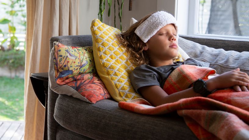 A young child lies on a couch under blankets with a flannel on their forehead.
