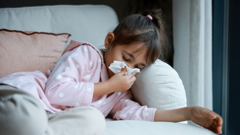 A little girl wipes her nose while resting on a couch.