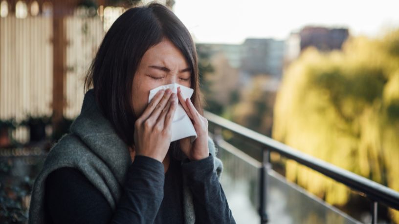 A woman stands on a balcony and coughs into a tissue.
