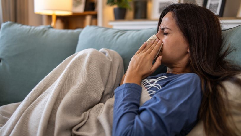 A woman lies on a couch under a blanket holding a tissue to her nose.