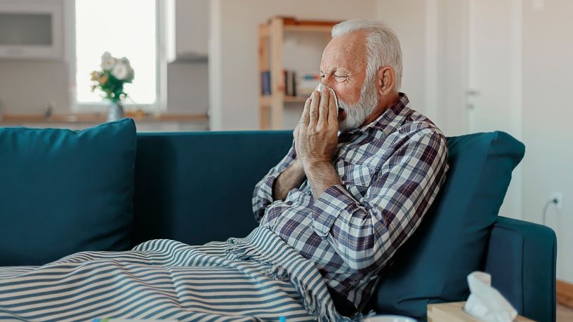 A man reclines on a couch and blows his nose with a tissue.