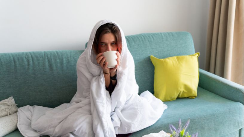 A young woman on the couch with a blanket over her head drinking a cup of tea.